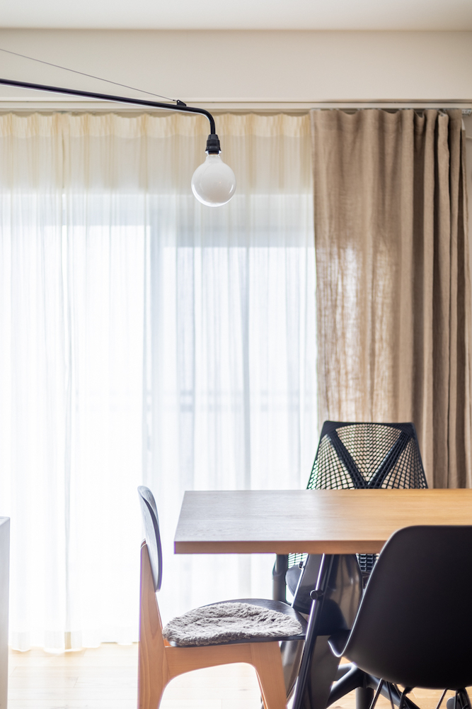 ダイニングのリノベーション事例。木製テーブルの上に配置された、黒いアームの可動式ブラケットライト(ウォールライト)。背景には大きな窓と、柔らかな質感のドレープカーテン