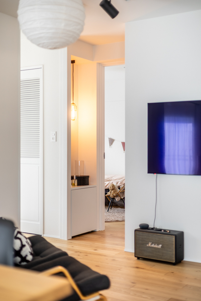 木の家具とペンダントライトが温かみを演出する、洗練されたモダンリビングのリノベーション事例。壁掛けテレビのあるリビングから、ガーランドが飾られた子ども部屋（寝室）が繋がる空間。
