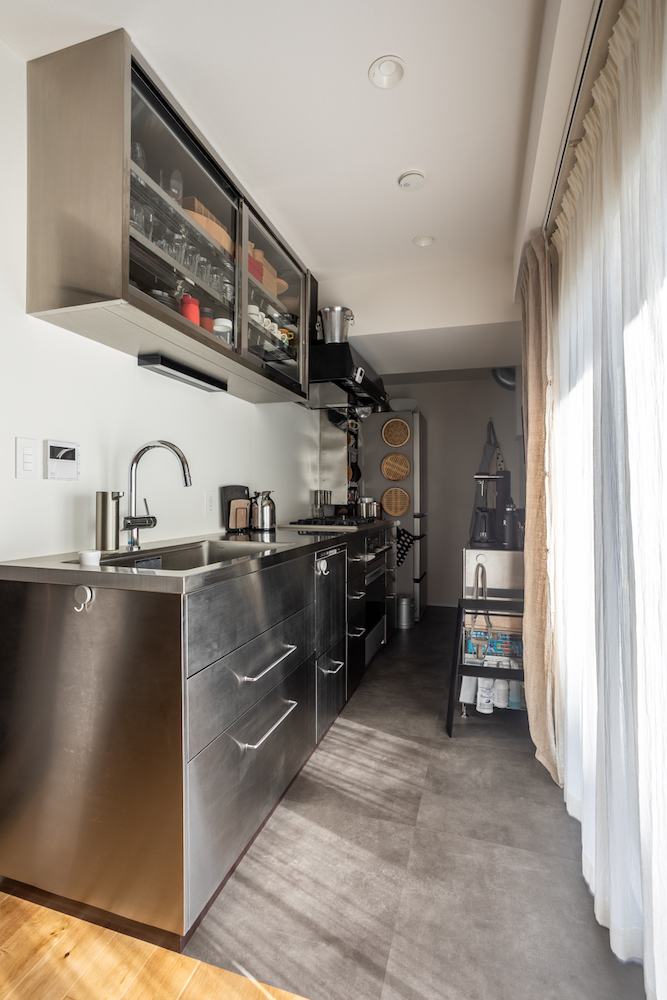 大きな窓から自然光が差し込む、壁付けオールステンレスキッチンのリノベーション事例。スタイリッシュな吊り戸棚と、手入れがしやすいグレーのフロアタイルを組み合わせた空間