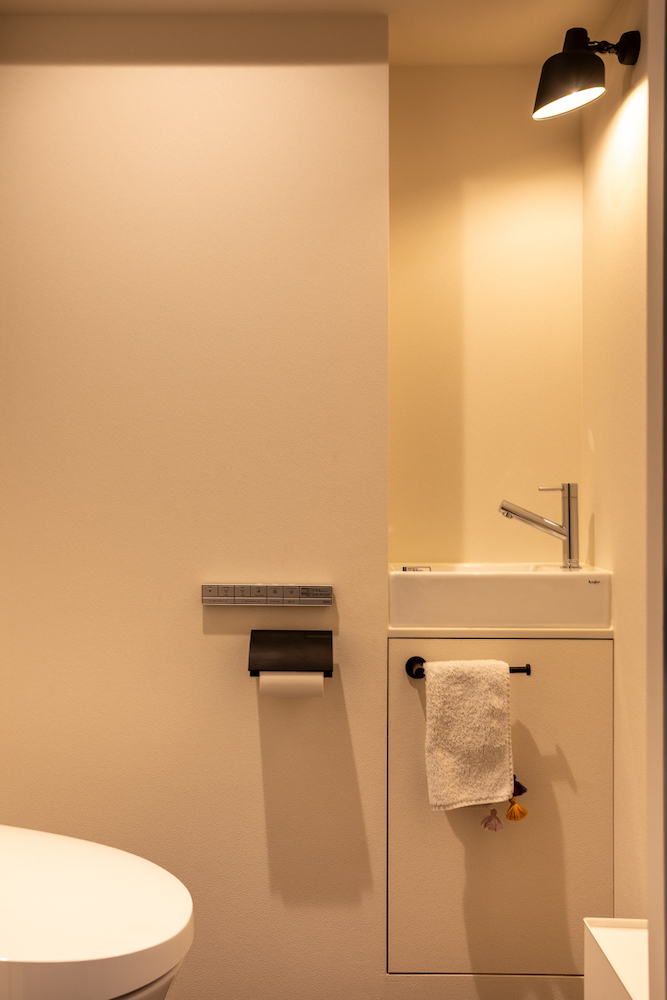 コンパクトな独立型手洗い器とブラケットライトを設けた、シンプルなトイレのリノベーション事例。マットな質感のペーパーホルダーやタオル掛けで統一された空間