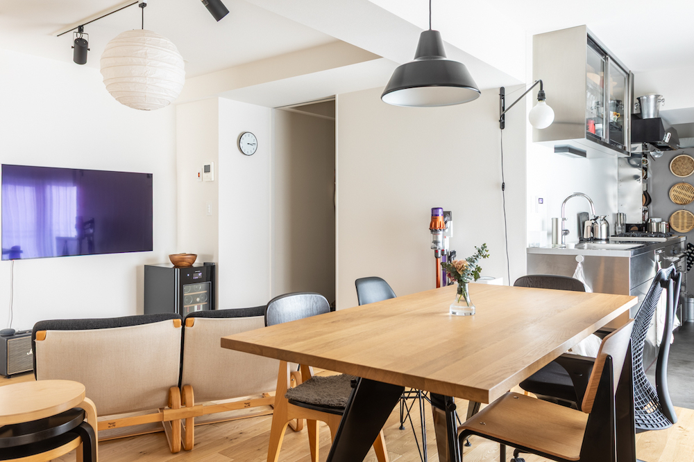 木の家具とステンレスキッチンを組み合わせた、開放的なLDK（リビング・ダイニング・キッチン）のリノベーション事例。壁掛けテレビのあるリビングからダイニングまでがシームレスに繋がる空間