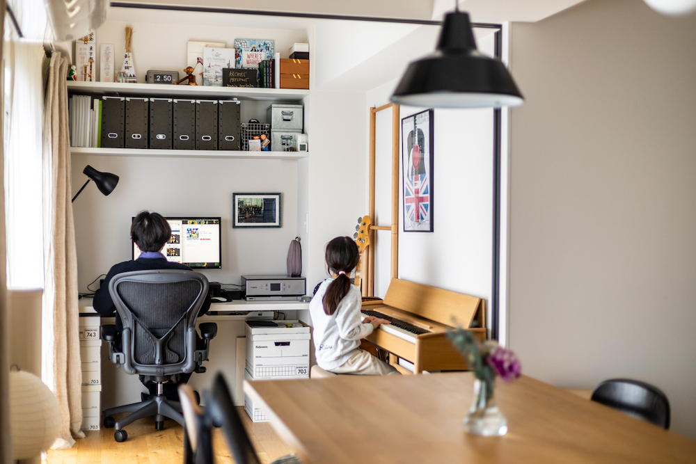 リビングの一角に造作したワークスペースのリノベーション事例。壁面収納付きのデスクでテレワークをする父親の隣で、子どもが木製ピアノを弾いている多目的空間