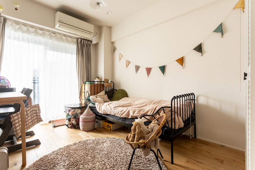 大きな窓から自然光が差し込む、明るい子ども部屋(キッズルーム)のリノベーション事例。黒いアイアンのデイベッドと、木製のデスク・チェアを配置。壁にはガーランドが飾られ、床には丸いラグと、ぬいぐるみが乗った子ども用チェアがある。フローリングは木の温もりを感じる
