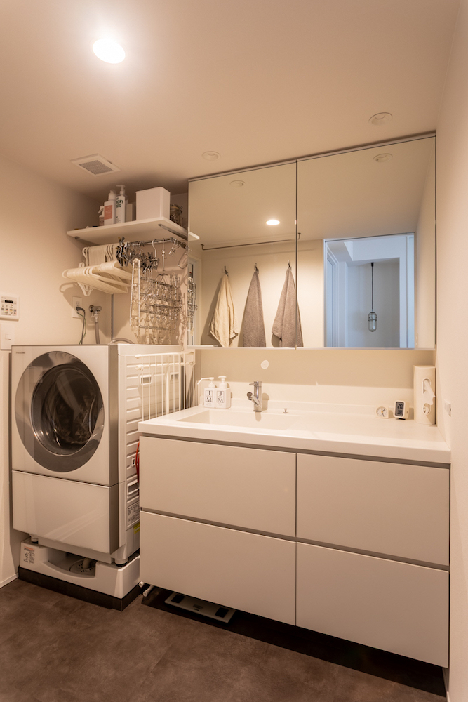 幅広の造作洗面台と大きな三面鏡を採用した、白基調の洗面脱衣室のリノベーション事例。洗濯機上には可動棚を設け、ハンガーや洗濯用品を機能的に収納。