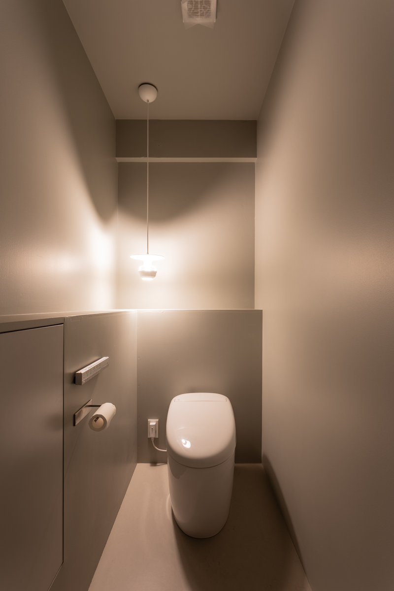 落ち着いたグレーの壁面と白いタンクレストイレがある、ミニマルなデザインのトイレスペース。左側のペンダントライトが温かい光を落としています。
